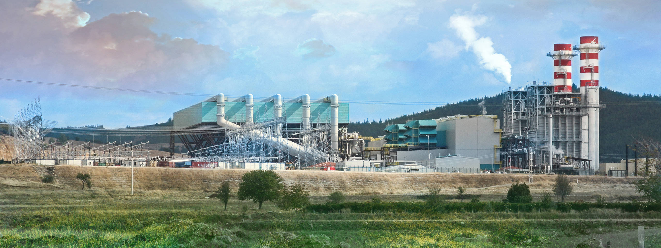 A large industrial power plant with cooling towers and high-voltage lines under a blue sky, surrounded by fields.