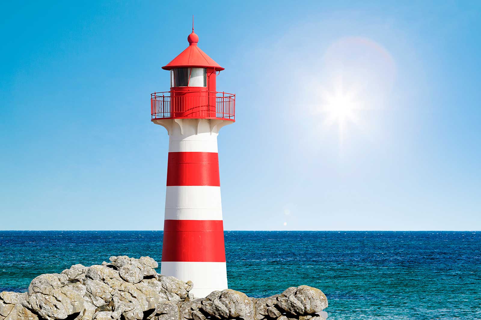 A red and white striped lighthouse stands on a rocky shore by a calm blue sea beneath a bright sun and clear sky.