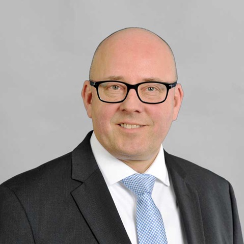A man in a suit with a blue tie, set against a grey background, appears professional. Portrait of Ingo Birnkraut.