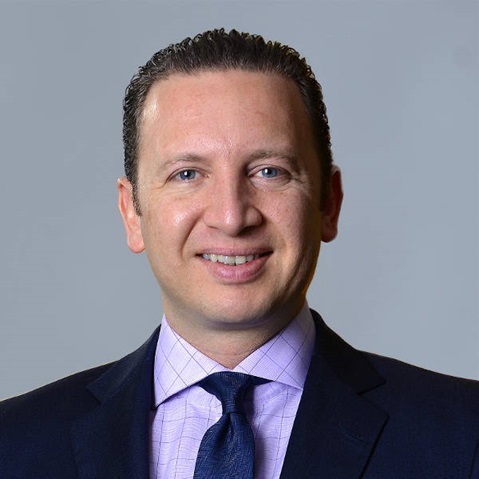 A professional man in a suit with a collared shirt and tie, set against a plain grey background.